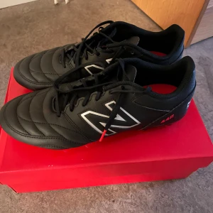 Svarta fotbollsskor från New Balance - Snygga svarta fotbollsskor från New Balance med snörning och vita detaljer. Skorna har en klassisk design med märkets logga på sidan och hälen. Perfekta för fotbollsplanen med en stilren look.