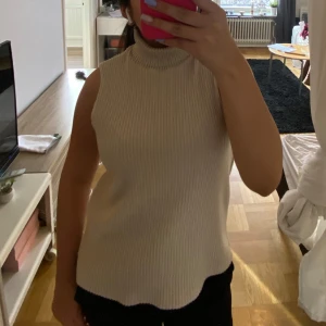 Beige polotopp - Säljer en stilren beige ribbad polotopp utan ärmar. Perfekt för lager-på-lager eller att bära som den är. Den har en hög krage som ger en elegant touch. Passar bra till både jeans och kjol.