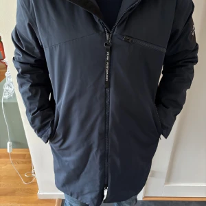 Mörkblå jacka från Peak Performance - Säljer en stilren mörkblå jacka från Peak Performance. Jackan har en dragkedja framtill och en praktisk huva. Den har en ficka med dragkedja på bröstet och en logotyp på ärmen. Perfekt för kyligare dagar.