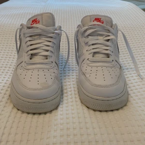 Vita Nike Air Force 1 sneakers - Säljer ett par klassiska vita Nike Air Force 1 sneakers med röd logga. Skorna har snörning och perforerade detaljer på tån för extra ventilation. Perfekta för en stilren look.