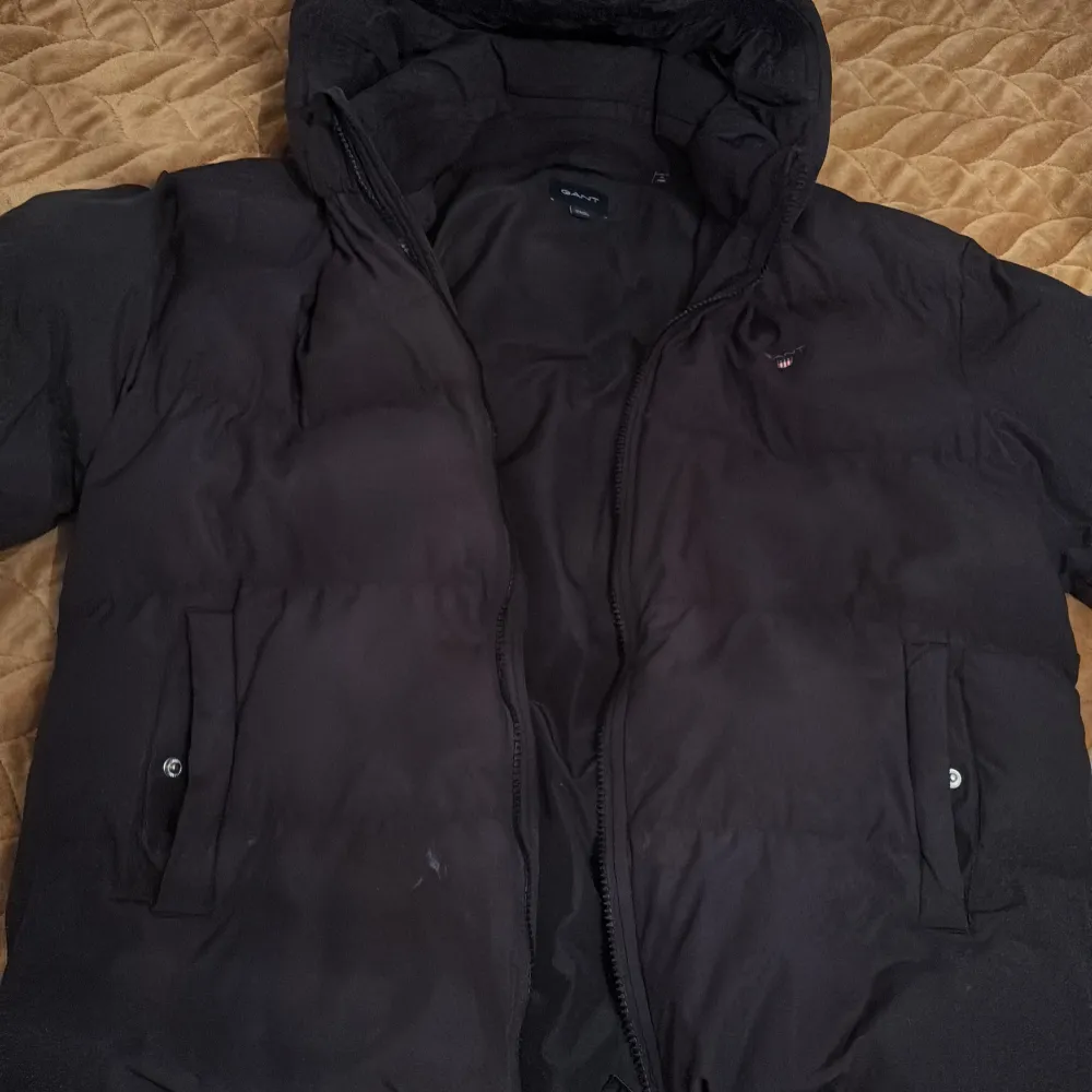 Säljer en svart dunjacka från GANT i storlek 176 cm. Jackan har en dragkedja framtill och praktiska fickor med knappar. Perfekt för kyliga dagar med sin vadderade design och huva.. Takit.