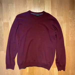 Vinröd merinoull tröja från Sand Copenhagen - Vinröd stickad tröja från Sand Copenhagen med rund halsringning och diskret broderad logga på bröstet. Klassisk modell med långa ärmar och ribbade muddar vid ärmslut och nederkant. Perfekt för lager-på-lager-stil.