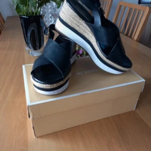 Svarta platåsandaler från Michael Kors - Nya ,bara provade .Säljer ett par snygga svarta sandaler från Michael Kors med platåsula och flätad detalj i naturfärgat material. Skorna har breda remmar, öppen tå och spänne vid ankeln. Perfekta för dig som vill ha både stil och höjd. Äkta läder.