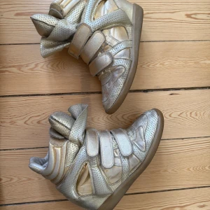 Guldiga Bekett sneakers Isabel Marant  - Säljer ett par speciella Bekett sneakers (såklart äkta!!!) från Isabel Marant i metallic guld med tre breda kardborreband och perforerade detaljer. Skickar gärna fler bilder i DM. Skorna har en 5 cm inbyggd kilklack och vadderad plös för extra statement-look. Perfekta för dig som vill sticka ut med din stil. Eftersom de är limited edition så är priset högre än de mer vanliga färgerna. De behöver putsas upp lite. Har tyvärr ingen låda kvar men kan skicka med en dustbag. 