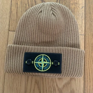 Beige ribbstickad mössa från Stone Island - Ribbstickad beige mössa från Stone Island med uppvikt kant och klassisk logotyp framtill. Märket är fastsytt med två knappar. Perfekt för kyliga dagar och enkel att matcha med olika outfits.