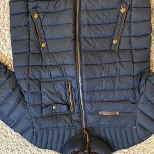 Mörkblå pufferjacka från Hansen & Jacob - Säljer en mörkblå pufferjacka från Hansen & Jacob med dragkedja och flera fickor med knappdetaljer. Jackan har ståkrage och quiltad design som ger en modern look. Perfekt för kyligare dagar.
