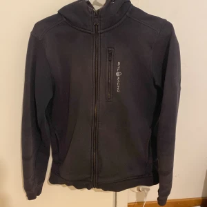 Svart zip-hoodie från Sail Racing - Svart hoodiejacka från Sail Racing med dragkedja och huva. Jackan har en bröstficka med dragkedja och Sail Racing-logga på bröstet samt tryck på huvan. Tillverkad i bomull och polyester för en mjuk känsla. KAN DISKUTERA PRISET 