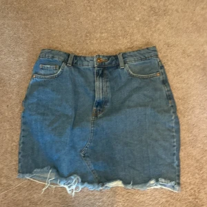Jeanskjol med fransig kant - Säljer en klassisk blå jeanskjol med fransig nederkant och femficksmodell. Kjolen har bälteshällor och knappstängning framtill. Perfekt för dig som gillar en enkel och tidlös stil.  Mindre i storlek 