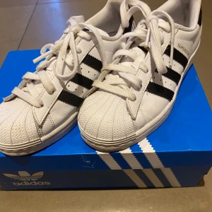 Adidas Superstar vita sneakers med svarta ränder - Klassiska Adidas Superstar sneakers i vitt med svarta ränder och tåskydd i gummi. Köpta på Plick och jag har själv aldrig haft på mig och den förra ägaren verkade ha haft på sig få gånger. 