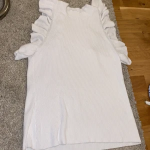 Vit ribbad topp med volangärmar - Säljer en vit ribbad topp med volangdetaljer längs ärmhålen. Toppen är ärmlös och har en mjuk, stretchig känsla. Perfekt för vår och sommar, enkel att matcha till olika outfits.