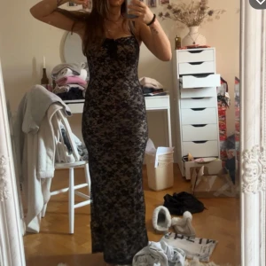 Svart spetsklänning med blommönster - Säljer en lång, tight svart klänning med blommig spets och smala axelband. Klänningen har en liten rosett framtill och är figurnära med transparent spets över ett beige foder. Perfekt för festliga tillfällen!