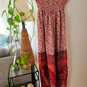 Röd mönstrad långklänning - Säljer en röd långklänning med bohemiskt mönster och smock upptill. Klänningen har en avslappnad passform och är perfekt för sommardagar. Färgerna går i olika röda och vita nyanser med paisley- och blommönster.