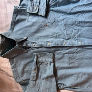 Ljusblå skjorta från Polo Ralph Lauren - Säljer en klassisk ljusblå skjorta från Polo Ralph Lauren med broderad logga på bröstet. Skjortan har lång ärm, knappar framtill och två bröstfickor med lock. Perfekt för en stilren look.