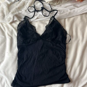 Svart spetslinne med smala axelband - Säljer ett svart linne med spetsdetaljer upptill och smala knytbara axelband. Djup v-ringning och mjukt material som känns bekvämt mot huden. Perfekt att styla till jeans eller kjol.