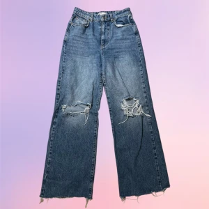 Blå slitna jeans med raka ben  - Säljer ett par blå jeans med raka, vida ben och slitna detaljer på båda knäna. Jeansen har hög midja och råa, fransiga benslut för en trendig look. Perfekta för dig som gillar en avslappnad och cool stil. Helt utan defekter