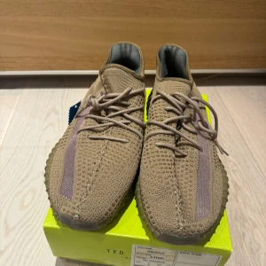 Adidas Yeezy Boost 350 V2 i beige - Säljer ett par Adidas Yeezy Boost 350 V2 sneakers i beige med lila inslag. Skorna har stickad ovandel, snörning och en ribbad sula i gummi. Perfekta för dig som gillar streetwear och vill ha något unikt på fötterna.