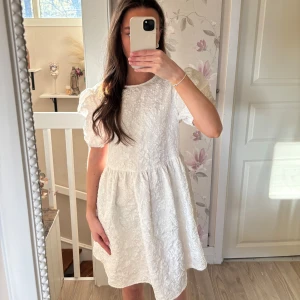 Studentklänning  - Säljer en söt vit kortklänning med puffärmar och rund halsringning. Klänningen har ett vackert strukturmönster i tyget och en lätt utställd kjol. Perfekt för sommarens alla tillfällen! Skulle passa en S också!