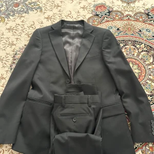 Svart kostym från Dressmann - Säljer en svart kostym från Dressmann i slim fit-modell, storlek 42/S/XS short. Kavajen har klassisk design med svarta knappar och röda detaljer på insidan. Byxorna är raka och matchar kavajen perfekt storlek 27W 30L. Har använt den 1 gång under balen och sens dess har bara suttit i garderoben alltså den är helt ny. Pris kan såklart diskuteras. OBS Skriv ner vad eran prisförslag är även om det blir litet så svarar jag troligtvis ja eftersom behlver pengarna just nu!