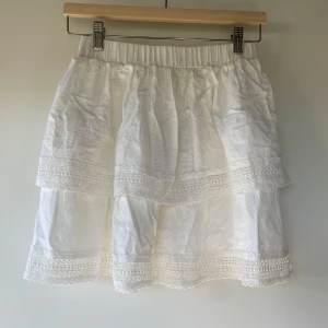 Vit volangkjol från Loavies - Superfin vit kjol från Loavies med dubbla volanger och spetsdetaljer. Kjolen har resår i midjan och är tillverkad i ett lätt och luftigt material, perfekt för varma dagar.