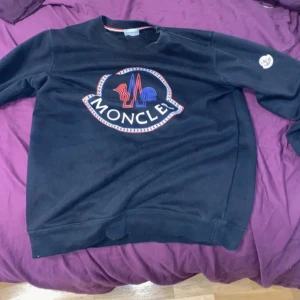 Svart sweatshirt från Moncler - Säljer en svart sweatshirt från Moncler med stort broderat Moncler-logo i rött, blått och vitt på bröstet. Tröjan har rund halsringning och lång ärm med en liten Moncler-patch på ena ärmen. Perfekt för en stilren och sportig look.