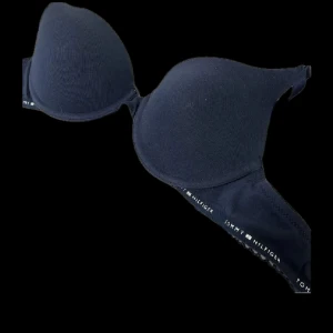 Mörkblå BH från Tommy Hilfiger - Mörkblå vadderad BH från Tommy Hilfiger med diskret logotyp längs bandet. Klassisk modell med justerbara axelband och hakknäppning bak. Mjuk och bekväm med slät yta och lätt rundade kupor.