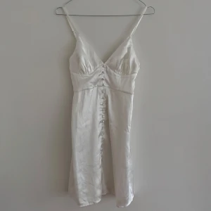 Vit satin klänning från Nelly - Säljer en elegant vit satin klänning från Nelly i storlek 34. Klänningen har smala axelband, knäppning framtill och en figurnära passform. Perfekt för studenten, midsommar eller andra sommar event! 