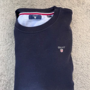 Mörkblå sweatshirt från GANT - Säljer en klassisk mörkblå sweatshirt från GANT med rund halsringning och liten broderad logotyp på bröstet. Tröjan har en enkel och stilren design med diskreta detaljer i rött och vitt på insidan av kragen.