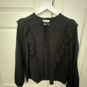 Svart blus med volang från KappAhl - Säljer en svart blus från KappAhl med volangdetaljer längs framsidan och axlarna. Blusen har långa ärmar och en liten knytning vid halsen. Perfekt för dig som gillar stilrena plagg med en twist. Pris går att diskutera och fråga om ni har en fråga!