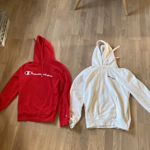 Champion hoodies i rött och vitt - Två snygga Champion hoodies, en röd i storlek small och en vit i storlek medium. Båda har huva, magficka och Champion-logga på bröstet. Perfekta för en avslappnad stil och tillverkade i mjukt bomullsmaterial.