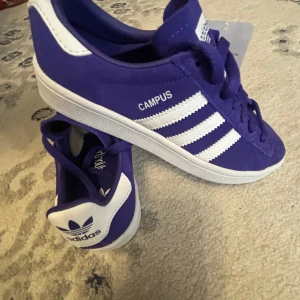 Lila Adidas Campus sneakers - Säljer ett par lila Adidas Campus sneakers med vita detaljer och klassiska tre ränder på sidan. Skorna har snörning och är i mocka med vit sula och vit hälkappa. Perfekta för dig som vill sticka ut med färg på fötterna!
