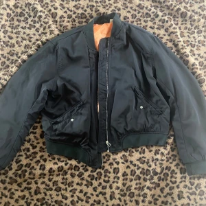 Svart bomberjacka med orange foder - Säljer en svart bomberjacka med klassisk passform och orange foder. Jackan har dragkedja framtill, två snedställda fickor med knappar och ribbade muddar vid ärmslut och midja. Perfekt för dig som vill ha en stilren och tidlös look.