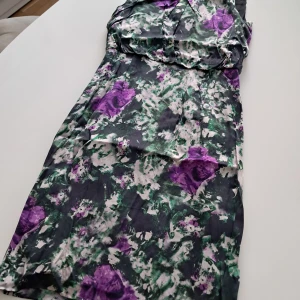 Blommig långklänning HUGO BOSS i siden. - Säljer en långklänning från HUGO BOSS med blommigt mönster i lila, grönt och vitt. Klänningen har en dekorerad krage med pärlor och svarta pärldetaljer. Materialet är siden, mjukt och glansigt, perfekt för dig som vill sticka ut.