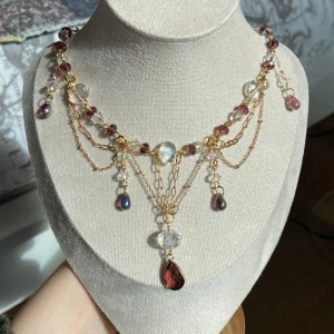 Guldhalsband med lila och klara pärlor - Unikt halsband med guldfärgade kedjor, lila och klara pärlor samt droppformade hängen. Halsbandet har flera lager kedjor och gnistrande detaljer som ger en lyxig känsla. Perfekt för att lyfta din outfit med elegans och färg.