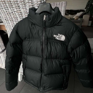 Svart dunjacka från The North Face - Säljer en svart dunjacka från The North Face med broderad logga på bröstet. Jackan har hög krage, dragkedja framtill och två sidofickor. Perfekt för kyliga dagar och riktigt skön att bära.