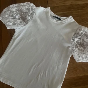 Topp Med Broderad Spets Från Zara - Superfin vit topp från Zara med korta puffärmar i transparent spets och broderade blommor. Nyskick, aldrig använd. Perfekt till våren och sommaren!
