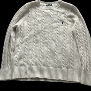 Vit kabelstickad tröja från Polo Ralph Lauren - Vit kabelstickad tröja från Polo Ralph Lauren med rund halsringning och broderad logga på bröstet. Tröjan är tillverkad i en blandning av ull, akryl och nylon och har långa ärmar samt ribbade muddar och nederkant.