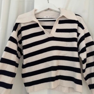 Randig stickad tröja från Mango - Säljer en krämvit och marinblå randig stickad tröja från Mango. Tröjan har v-ringning, stor krage och långa ärmar. Ribbad struktur och avslappnad passform, perfekt för en trendig vardagslook.💙🤍💙🤍