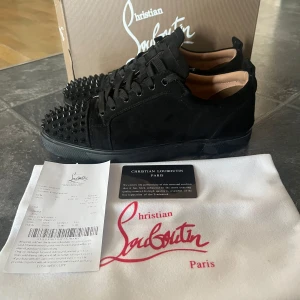 Svarta sneakers från Christian Louboutin - Säljer ett par svarta sneakers från Christian Louboutin med mattsvarta nitar på tån och sidorna. Skorna har klassisk röd sula, snörning och är tillverkade i mocka. Kommer med originalkartong, den är lite trasig däremot. Dustbag och kvitto.
