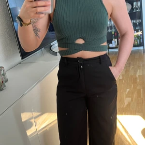 Grön ribbad croppad topp med cut-out detaljer - Snygg grön ribbad croppad topp med hög hals och unika cut-out detaljer framtill och i midjan. Toppen är ärmlös och har en tight passform som framhäver figuren. Perfekt för dig som vill sticka ut med en trendig look.