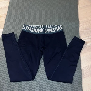 Svarta tränings tights från Gymshark - Säljer ett par mörkblåa tights från Gymshark med bred resår i midjan där loggan syns tydligt i vitt. Tightsen har en tight passform, är midwaist och är perfekta för träning. Materialet är stretchigt och bekvämt. Knappt använda.
