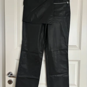 Svarta byxor med dragkedjedetalj - Unika svarta byxor med glansig finish och en stor asymmetrisk ficka framtill. Byxorna har en synlig silverfärgad dragkedja och raka ben. Perfekt för dig som vill sticka ut med en cool och modern look.