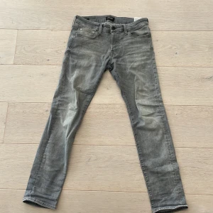Grå slim fit jeans från Jack & Jones - Säljer ett par gråa slim fit jeans från Jack & Jones, modell Glenn. Jeansen har klassisk femficksdesign, dragkedjegylf och snygga slitningar framtill för en cool look.