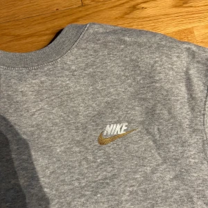 Grå sweatshirt från Nike - Säljer en klassisk grå sweatshirt från Nike med deras ikoniska logga i vitt och guld på bröstet. Tröjan har långa ärmar och en rund halsringning. Perfekt för en avslappnad stil. Inga tecken på användning, strl S