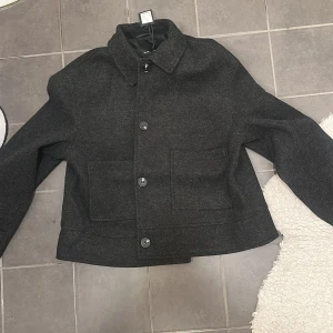 Mörkgrå kappa  med knappar - Säljer en stilren mörkgrå jacka med knappar framtill. Jackan har en klassisk krage och två fickor på framsidan. Perfekt för kyligare dagar och ger en snygg look. Passar bra till både jeans och chinos.