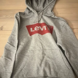 Grå hoodie från Levi's - Säljer en klassisk grå hoodie från Levi's med röd logga på bröstet. Tröjan har en stor ficka framtill och en bekväm huva med dragsko. Perfekt för en avslappnad stil. Supersnygg💕