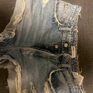 Blå jeansshorts  - Säljer ett par coola blå jeansshorts från Zara med fransiga detaljer och en avslappnad look. Perfekta för sommardagar och festivaler. De har en klassisk femficksdesign och knappar framtill. Medel midja
