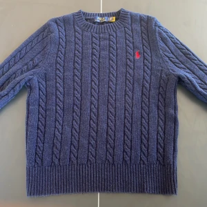 Mörkblå stickad tröja från Polo Ralph Lauren - Säljer en snygg mörkblå stickad tröja från Polo Ralph Lauren med kabelstickat mönster och röd broderad logga på bröstet. Perfekt för kyliga dagar med sin klassiska design och bekväma passform. 