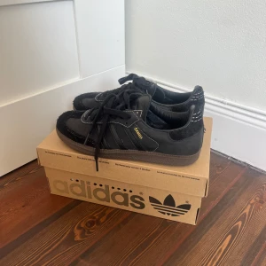 Svarta Adidas Samba OG x Ssense - Snygga svarta Adidas Samba OG sneakers med klassisk design. Aldrig använda. Skorna har en överdel i mocka och syntet med snörning och den ikoniska tre-randiga loggan på sidan. Perfekta för en stilren look. Köpta på Ssense.