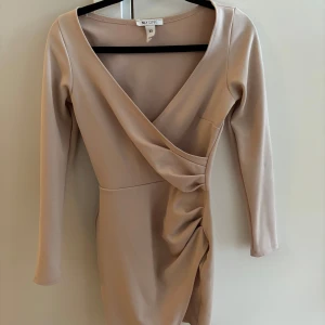 Beige omlottklänning från NLY One - Snygg beige omlottklänning från NLY One i en mjuk polyesterblandning. Klänningen har långa ärmar och en v-ringad design med rynkdetaljer på sidan. Perfekt för en stilren look.