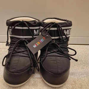 Svarta Moon Boots - Snygga svarta Moon Boots med vit sula och logotypdetaljer. De har snörning framtill och är perfekta för kalla vinterdagar. Den vadderade insidan ger extra värme och komfort. Aldrig använda. 